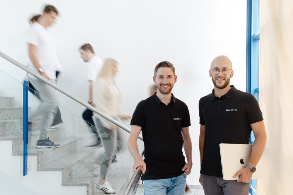 digital ZEIT Team Karriere Treppenhaus Zwei Mitarbeiter von digital ZEIT im Treppenhaus – dynamisches Team mit Laptop und digital ZEIT Poloshirts