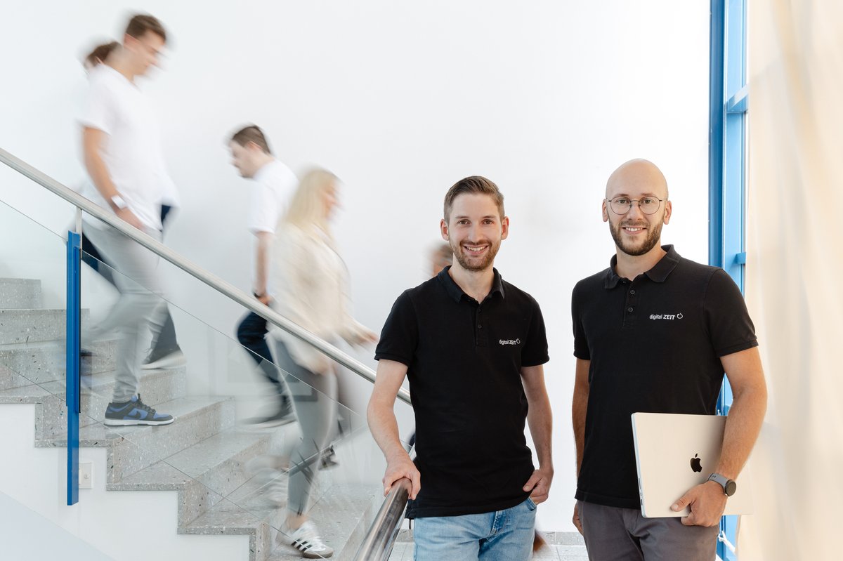 Zwei Mitarbeiter von digital ZEIT im Treppenhaus – dynamisches Team mit Laptop und digital ZEIT Poloshirts
