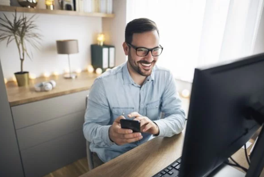 Mann im Homeoffice vor einem PC mit Smartphone in der Hand