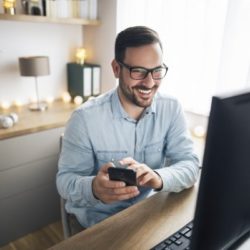 Mann im Home Office vor PC mit Smartphone in der Hand